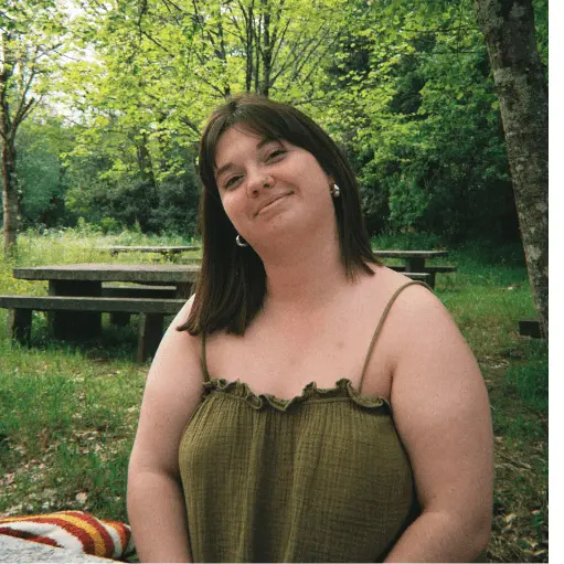 Fernanda Souza. Mulher sorridente sentada em um parque, usando vestido verde com alças finas, cercada por árvores e mesas de piquenique ao fundo.